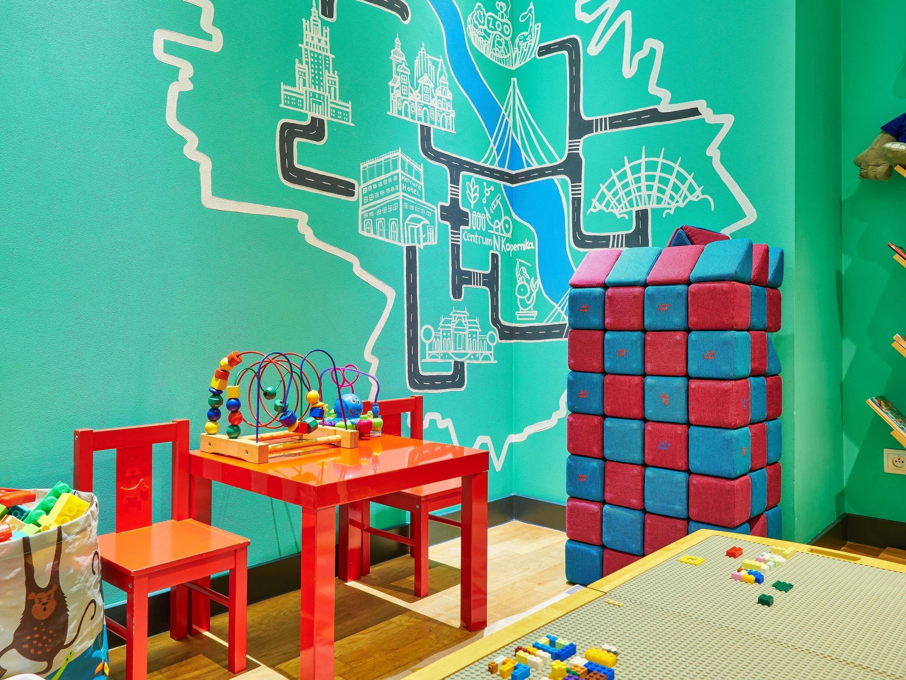 Zona de juegos infantiles con un mural colorido, una mesa roja y una estructura de bloques de juguete.