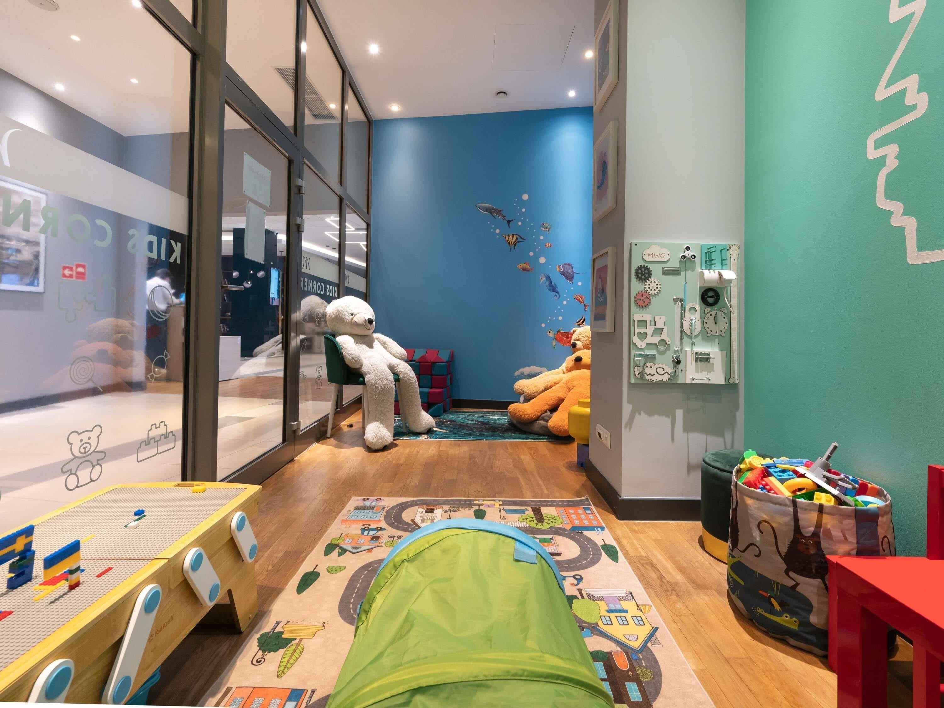 A children's playroom with a large teddy bear, a green tent, and a play mat with a cityscape design.