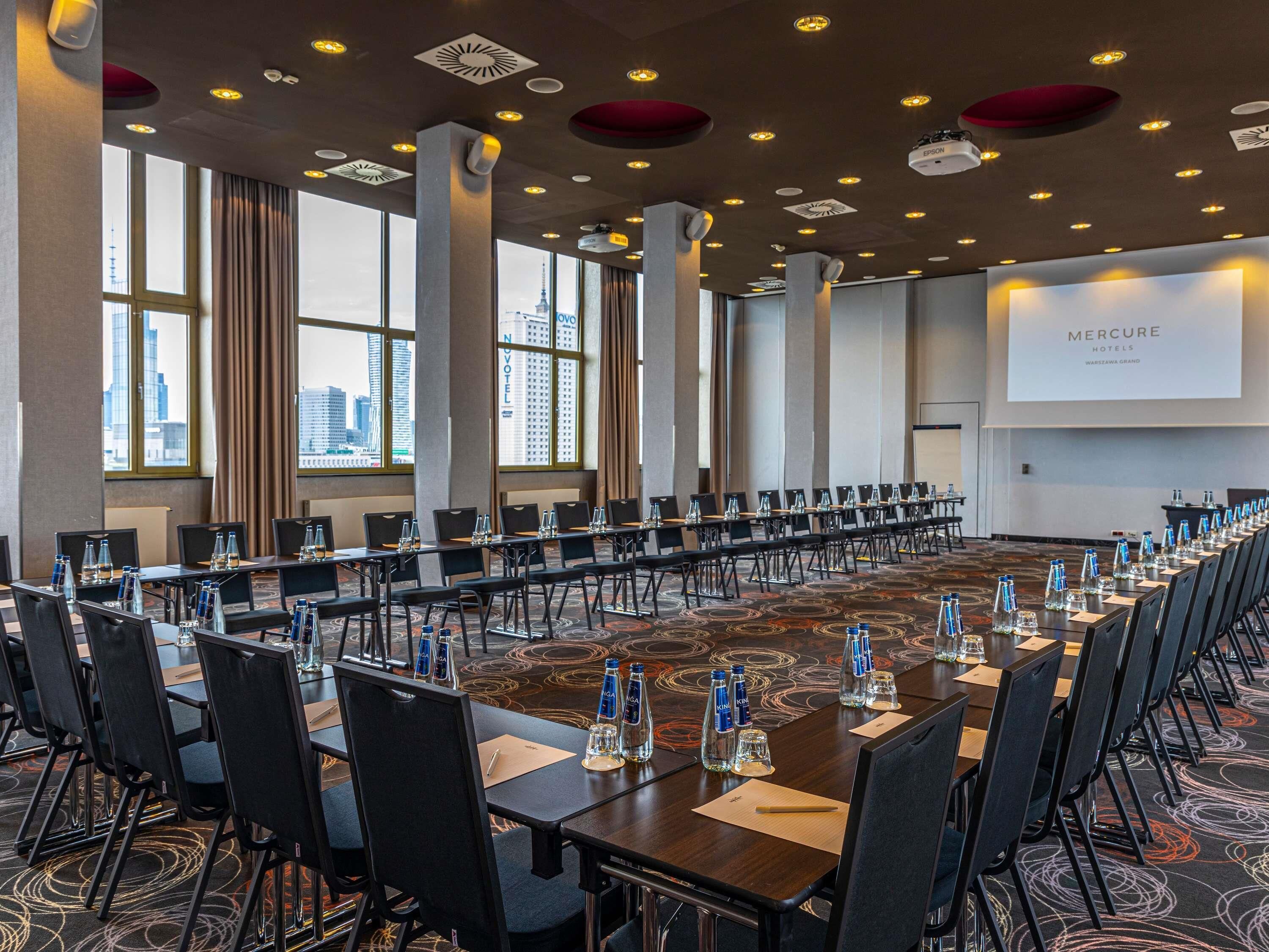 Konferenssitila, jossa on pitkiä pöytiä, tuoleja, vesipulloja ja projektorikangas, jolla näkyy teksti ”Mercure Hotels Warsaw Grand”.