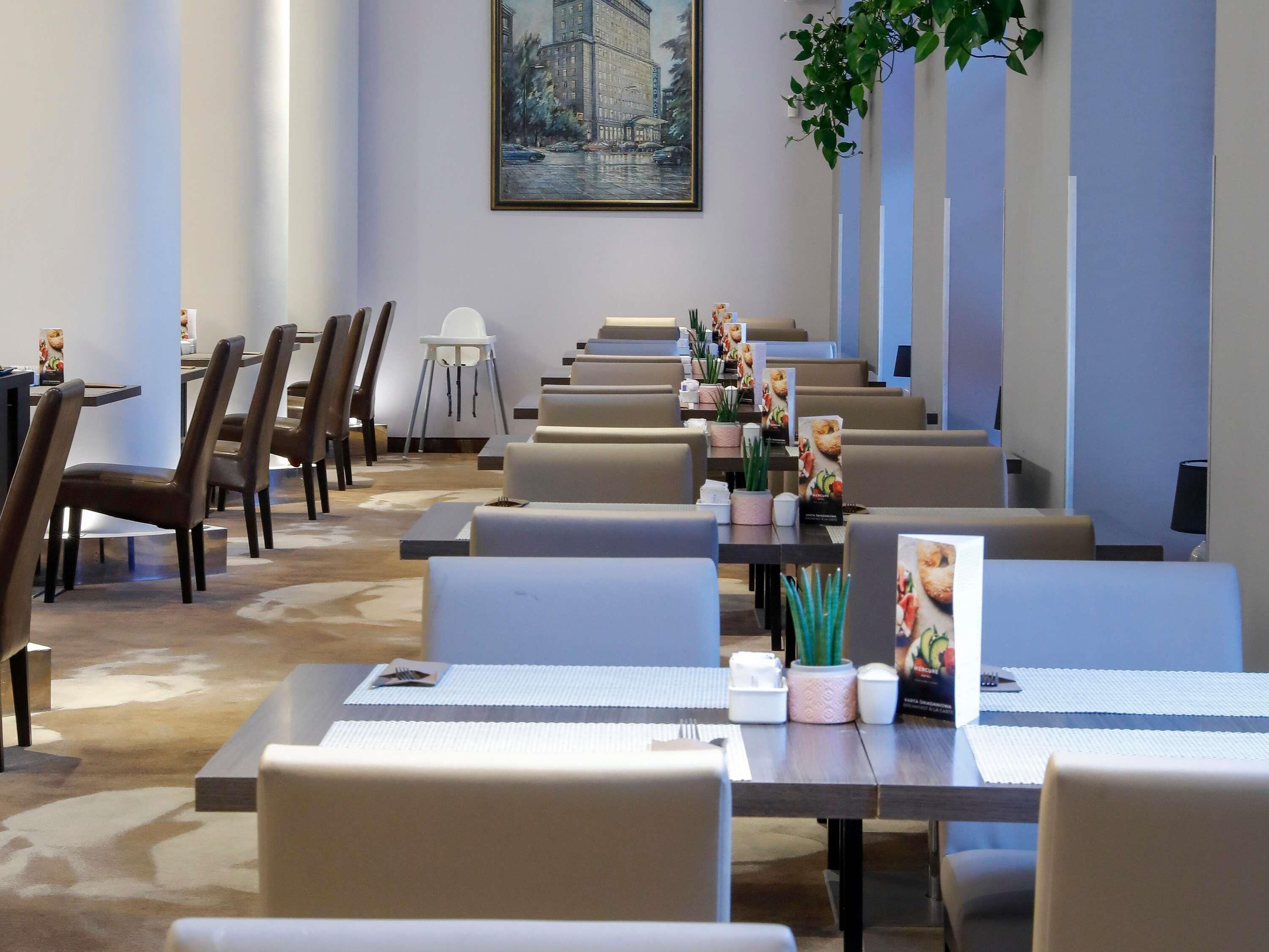A restaurant with long tables and chairs, a painting on the wall, and potted plants.