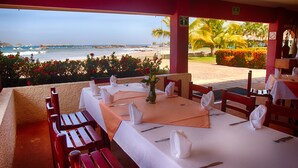 A restaurant with a table set for two, overlooking a beach with palm trees and flowers. - Qualton Club Ixtapa - All Inclusive (Ixtapa)