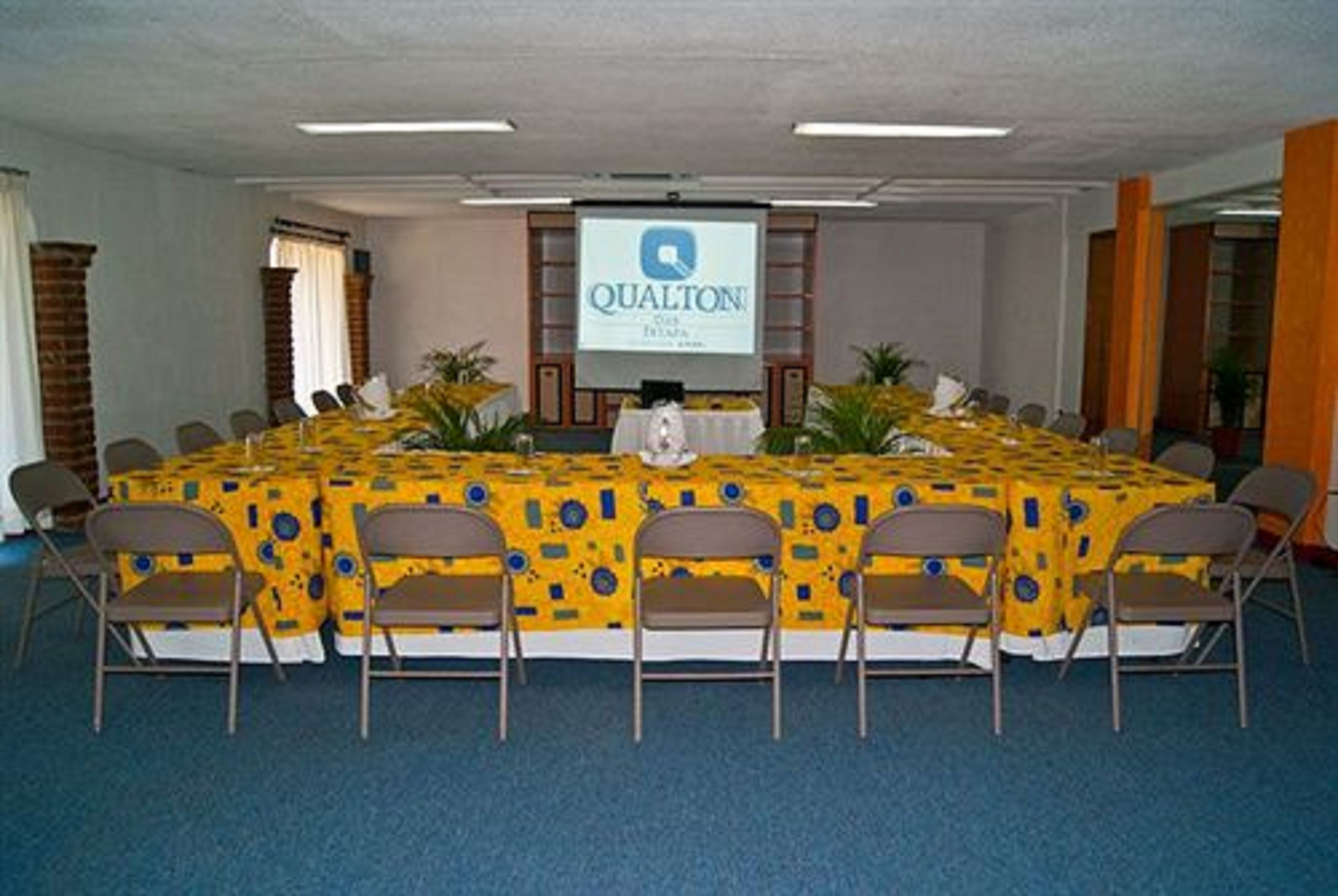 A conference room with a large table covered with a yellow tablecloth, surrounded by chairs. A screen displaying the "QUALTON" logo is mounted on the wall.