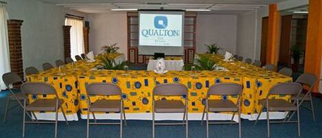 A conference room with a large table covered with a yellow tablecloth, surrounded by chairs. A screen displaying the "QUALTON" logo is mounted on the wall.