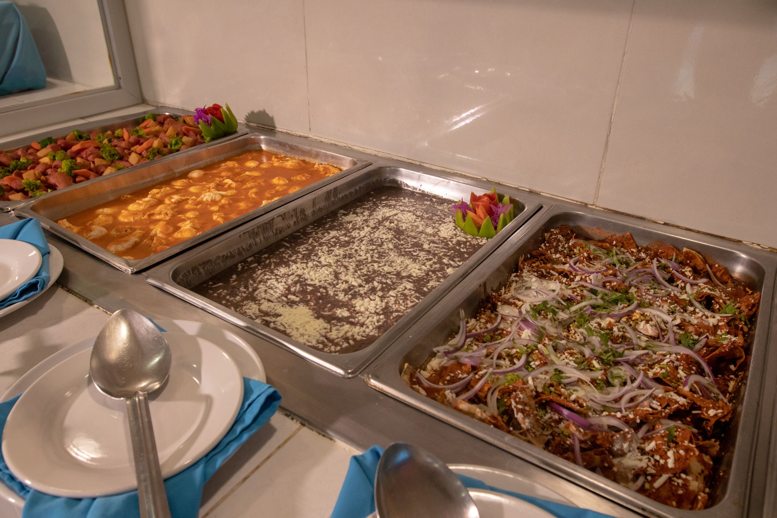 Un bufé con diversos platos, incluyendo una bandeja de enchiladas, una bandeja de arroz y una bandeja de verduras mixtas.