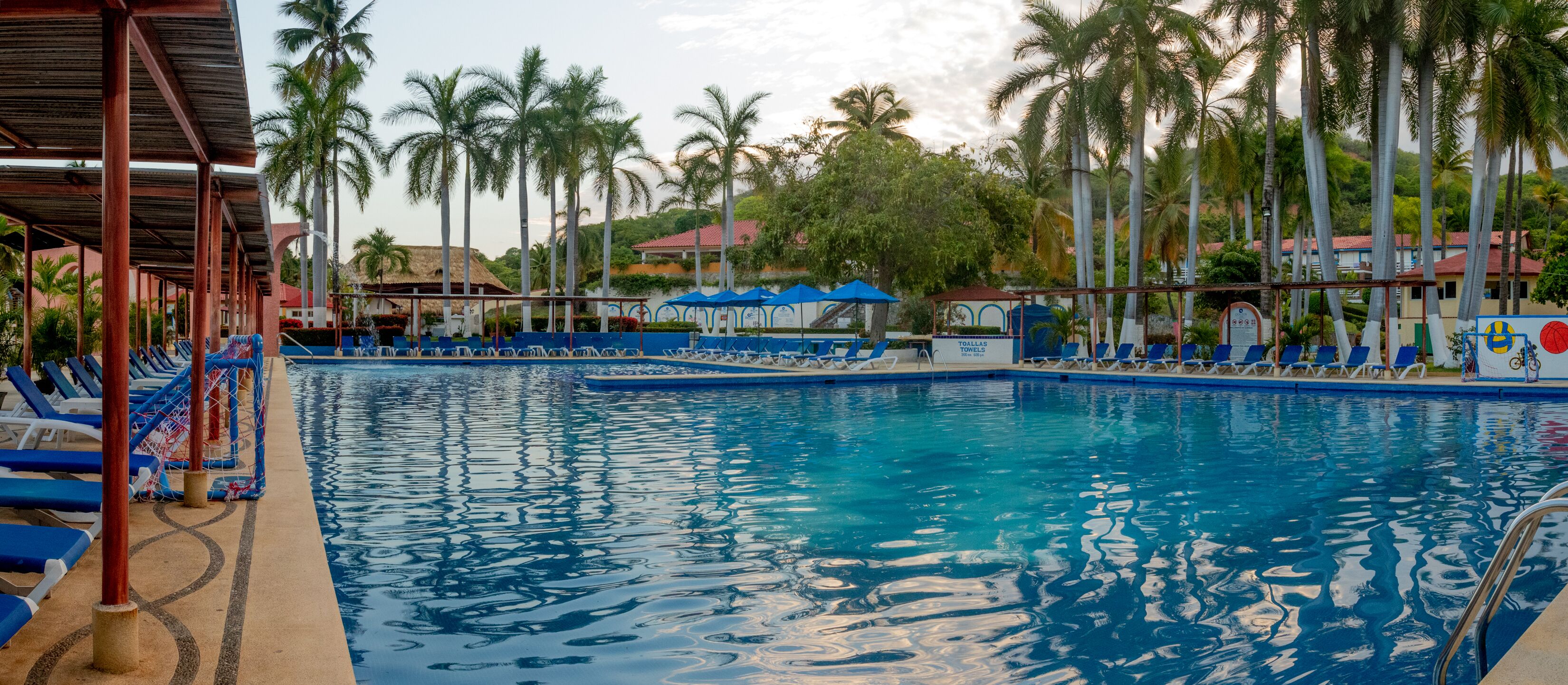 A swimming pool with blue and white lounge chairs, palm trees, and a covered area with a roof.