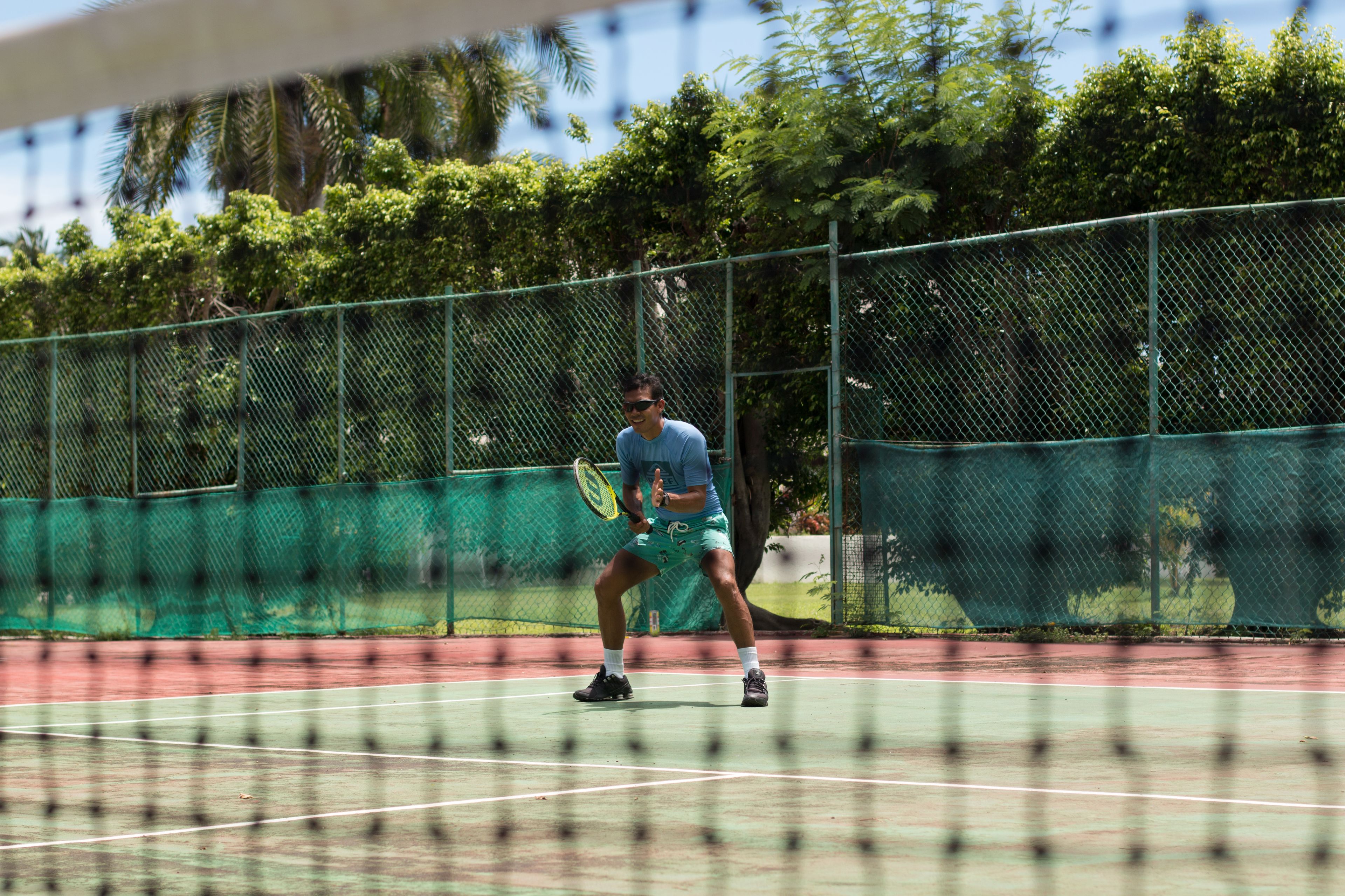 Ein Tennisplatz mit Netz, umgeben von einem grünen Zaun und Bäumen.