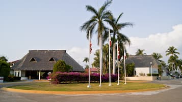 Un resort con edificios de techo de paja, palmeras y un césped cuidado con flores moradas.