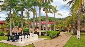 A chessboard set outdoors surrounded by palm trees and a pathway leading to a building with a red roof. - Qualton Club Ixtapa - All Inclusive (Ixtapa)
