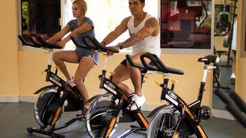 Dos personas pedaleando en bicicletas estáticas en un gimnasio con espejos y equipamiento de ejercicio.