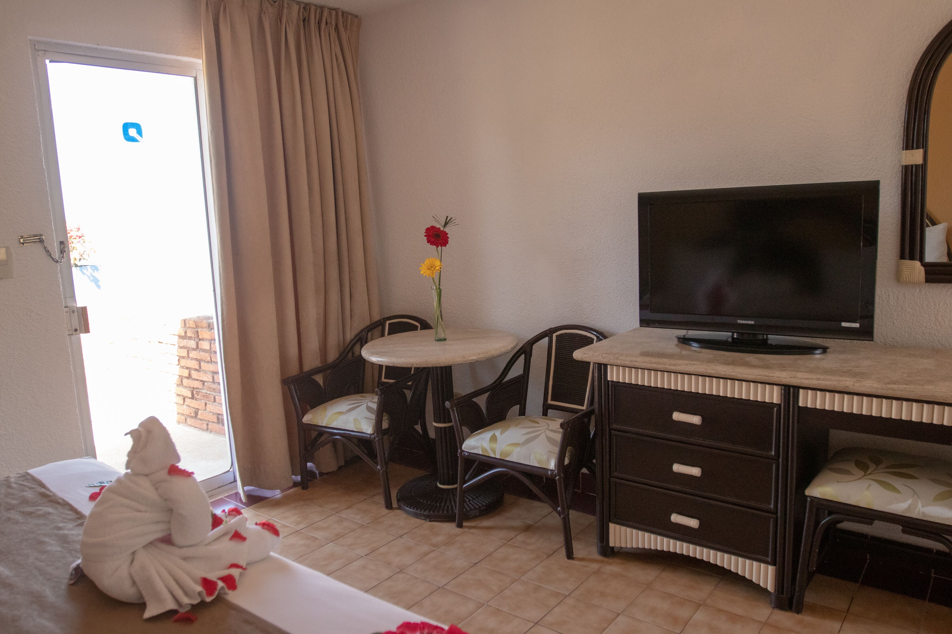 A hotel room with a bed, a TV, a small round table with chairs, and a window with curtains.
