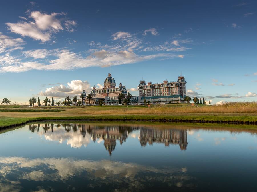 A large, historic building with a reflection in a calm body of water, surrounded by open grassy areas and a clear sky.