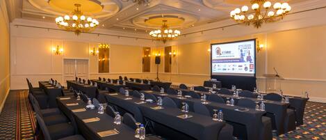 A conference room with rows of tables, chairs, and a projector screen displaying a presentation.