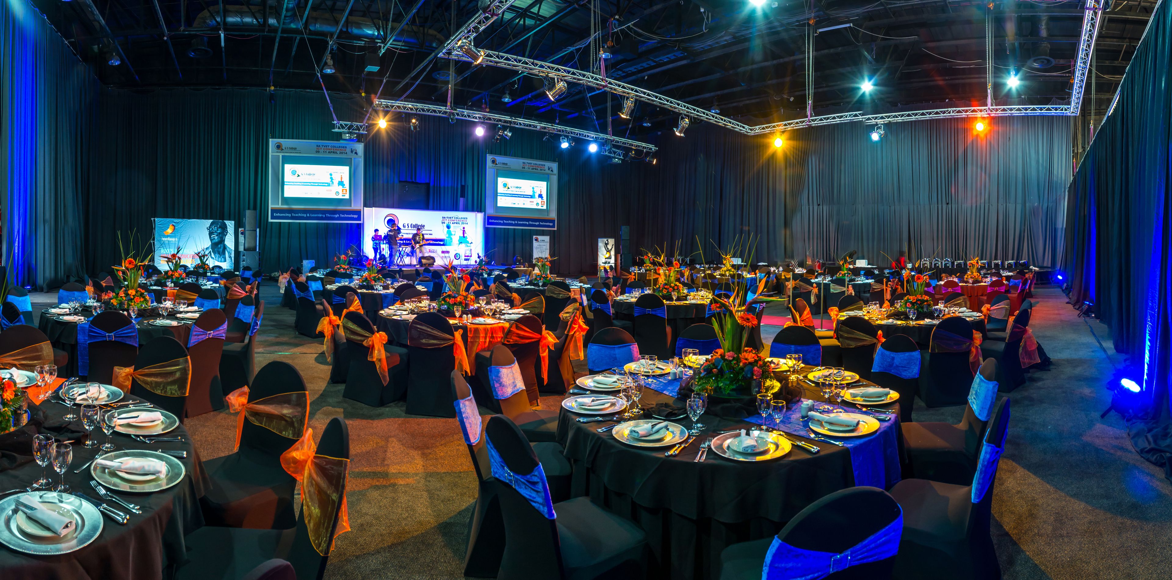 Een feestzaal met ronde tafels gedekt voor een formeel evenement, met een podium, een presentatiescherm en een band.