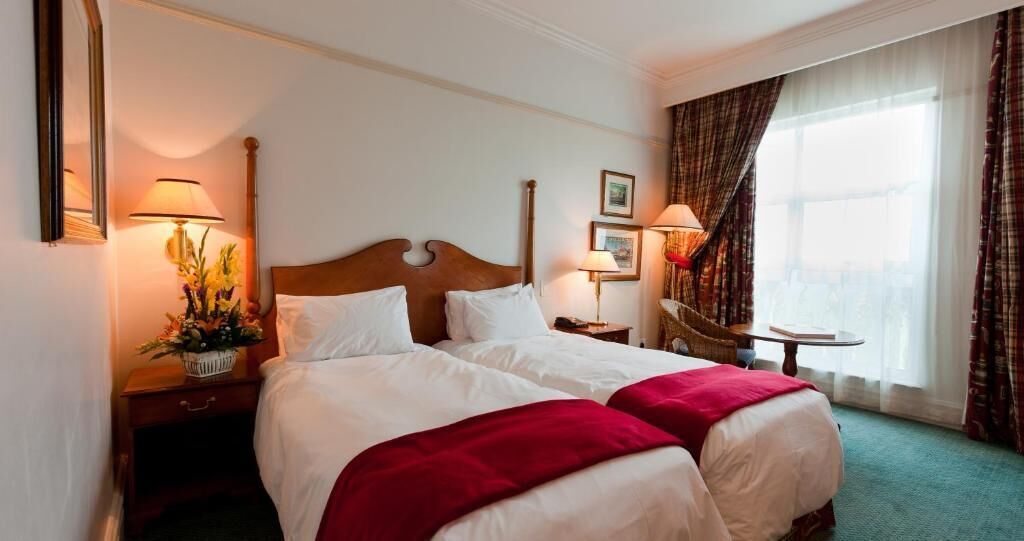 A hotel room with two beds, a wooden headboard, a nightstand with a lamp, a small table, and a window with curtains.