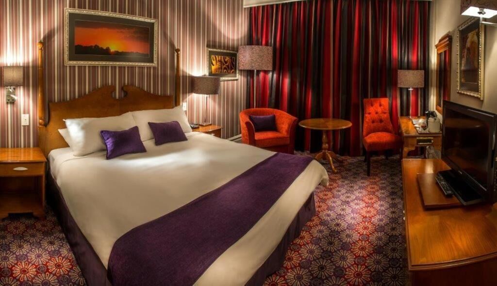 A hotel room with a large bed, two red armchairs, a small round table, a TV, and a fireplace.