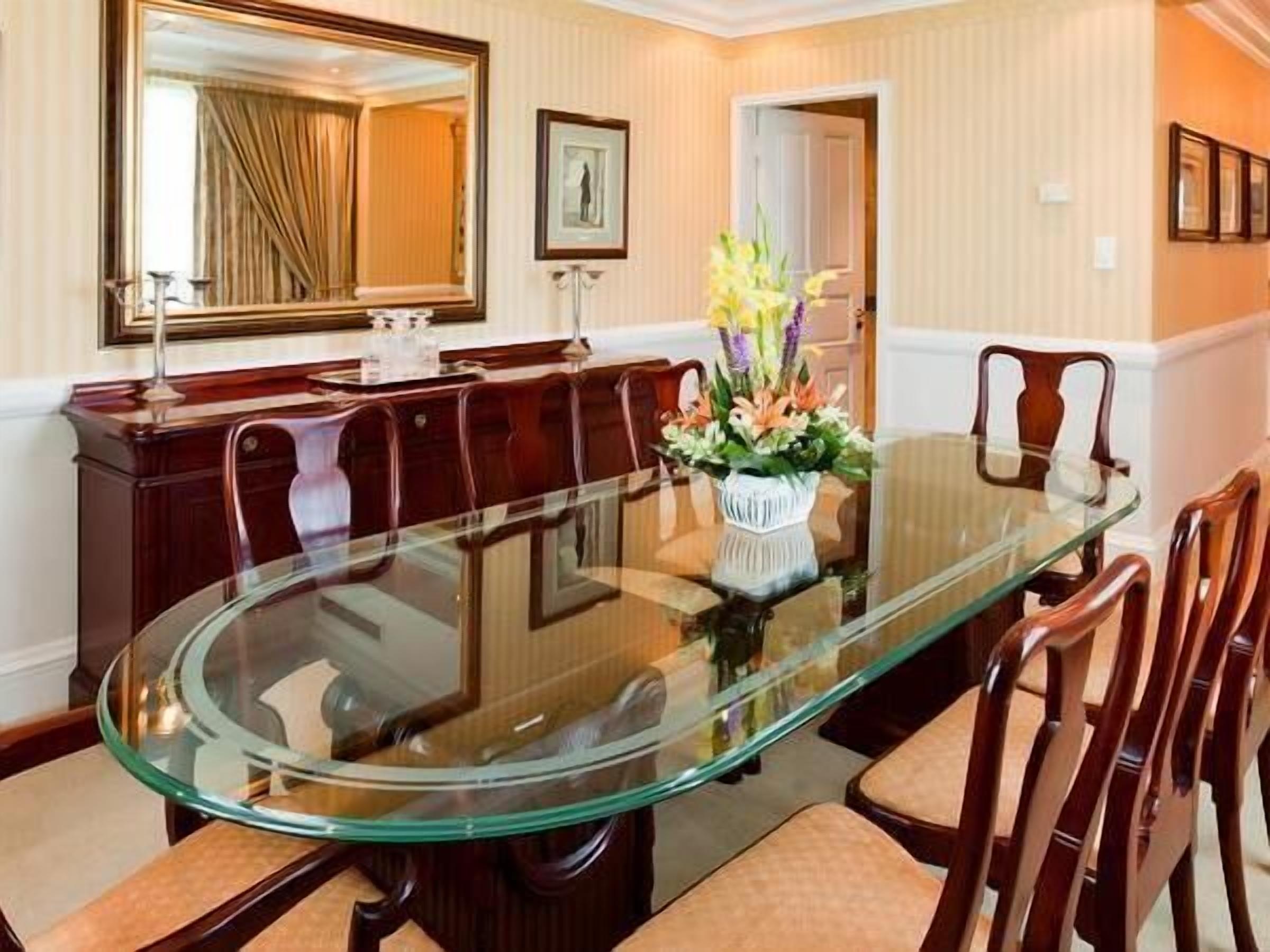 A dining area with a glass-top table, wooden chairs, a vase with flowers, a mirror, and a framed picture on the wall.