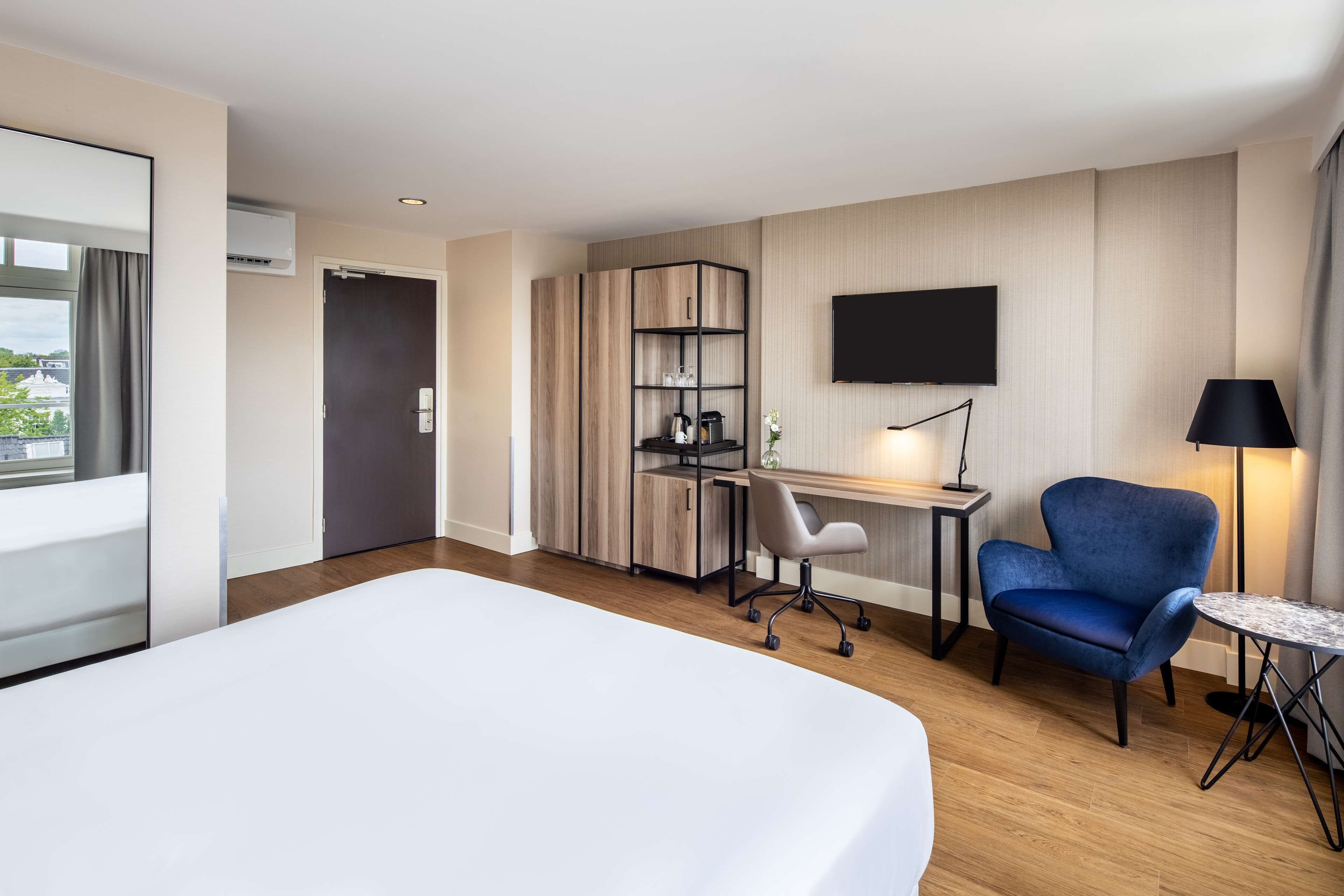 A hotel room with a large bed, a desk with a chair, a TV mounted on the wall, and a small table with a lamp.