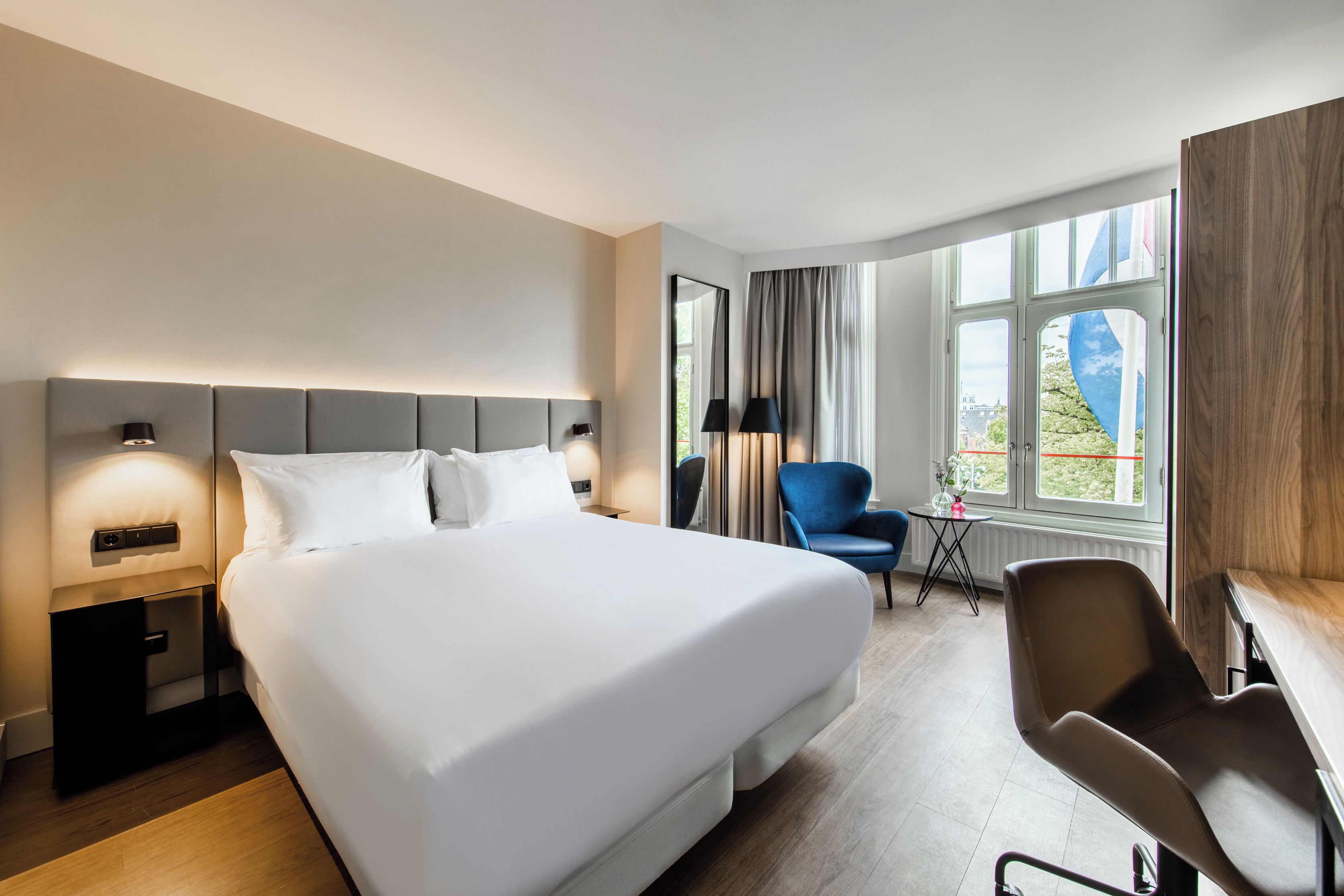 Habitación de hotel con una cama grande, un escritorio, una silla, una mesita de noche y una ventana con vistas.
