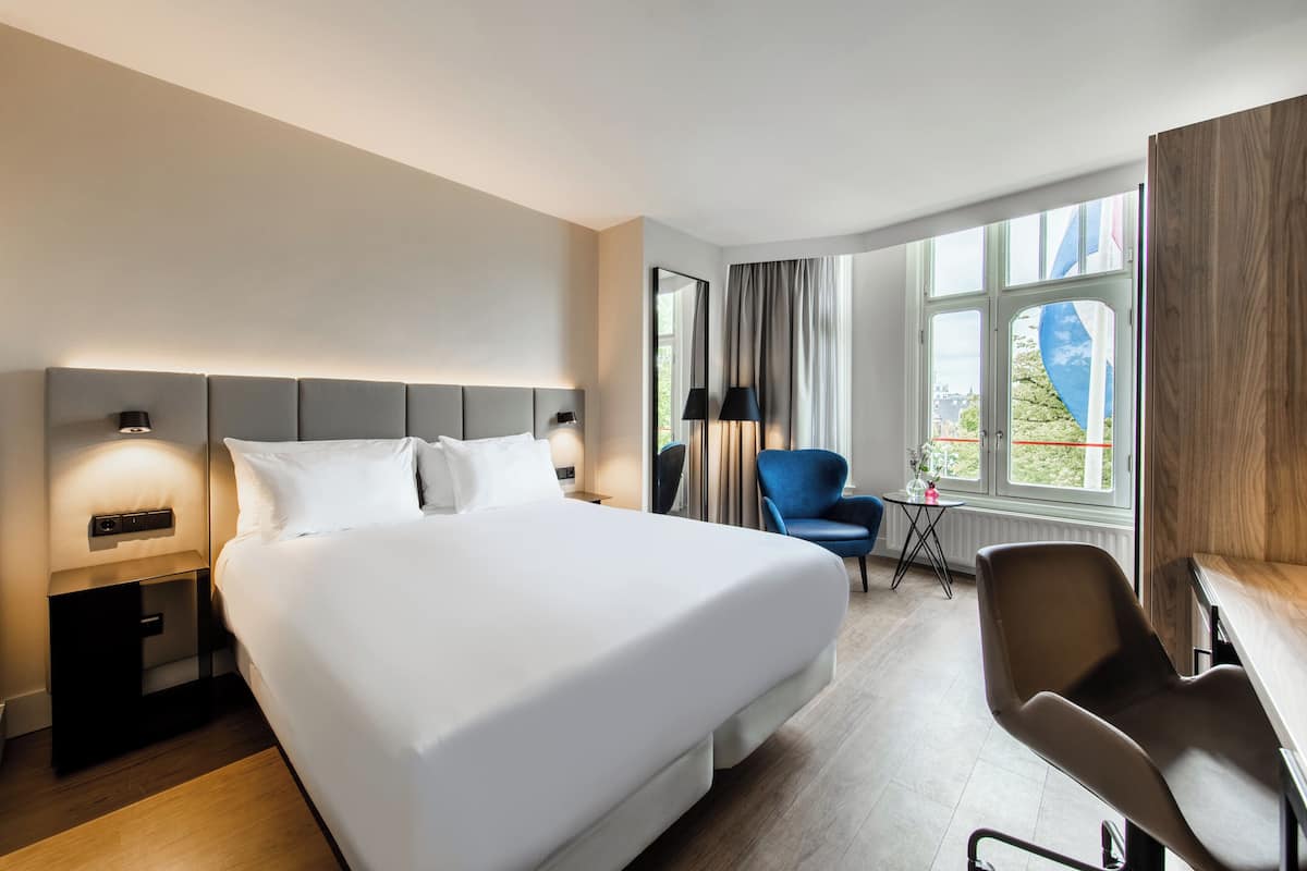Habitación de hotel con una cama grande, un escritorio, una silla, una mesita de noche y una ventana con vistas.