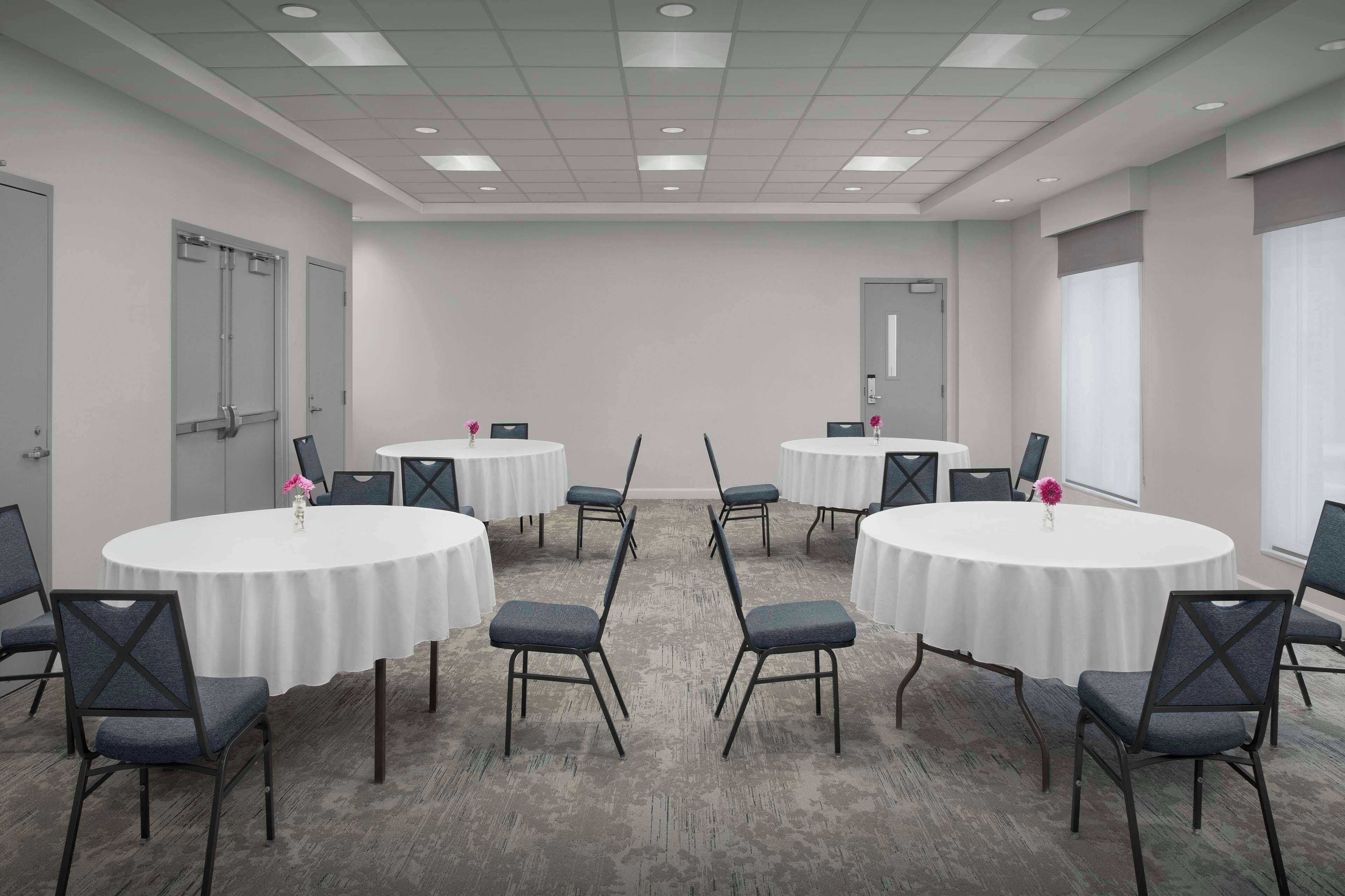 A conference room with four round tables, each surrounded by six chairs. The room has a carpeted floor and a plain wall with a door and a window.