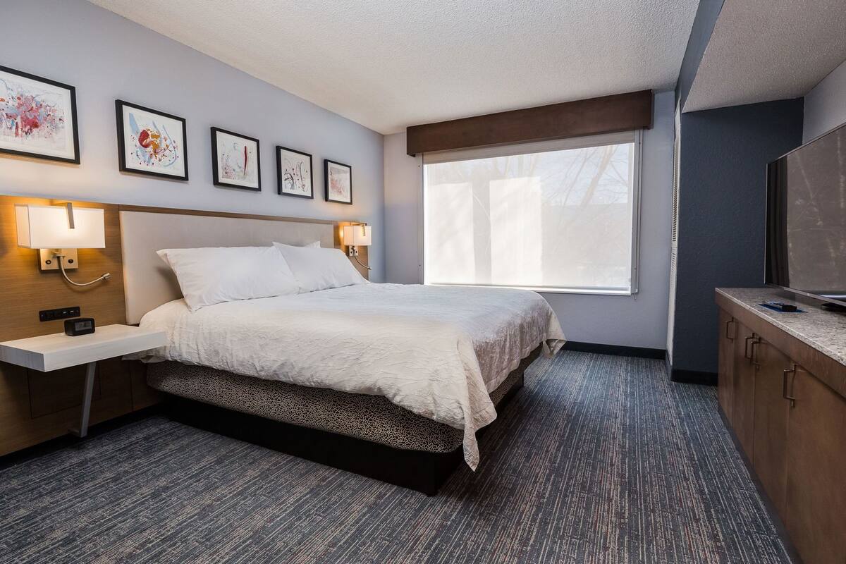 A hotel room with a bed, bedside lamps, a TV, artwork on the wall, and a window.