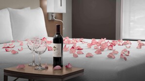 Una cama con pétalos de rosa, una botella de vino y dos copas sobre una mesita de noche en una habitación de hotel.