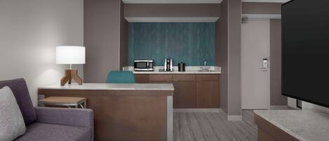 Habitación de hotel compacta con kitchenette, un sofá, un televisor y una pequeña mesa con una lámpara.