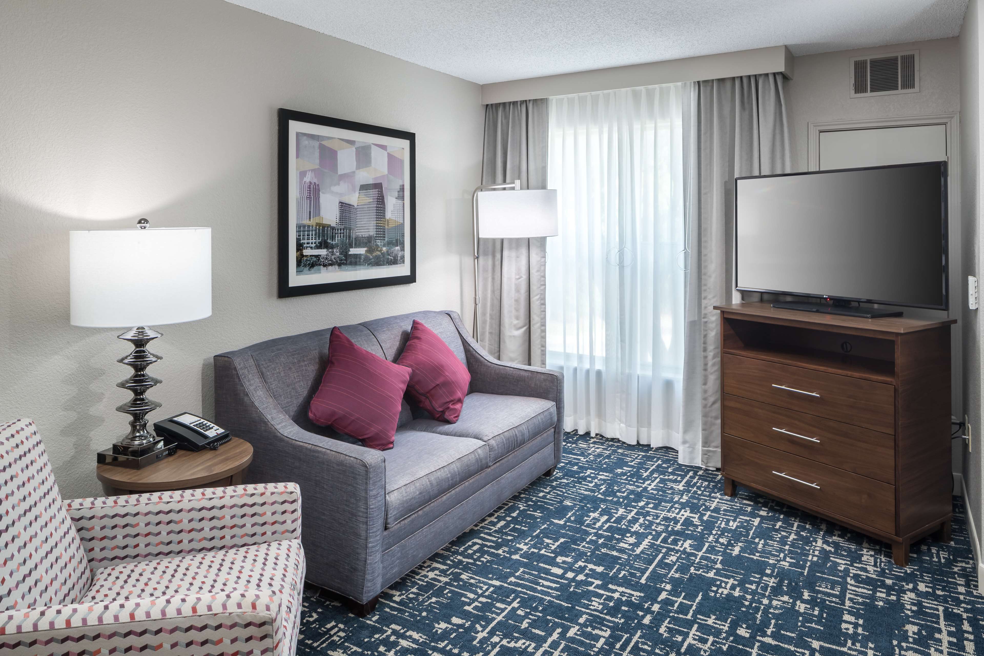 A hotel room with a grey sofa, a patterned armchair, a wooden cabinet, a flat-screen TV, a lamp, and a framed cityscape picture on the wall.
