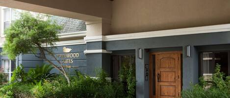 A building entrance with a wooden door, a sign reading "Homewood Suites," and a brick pathway leading to the entrance.