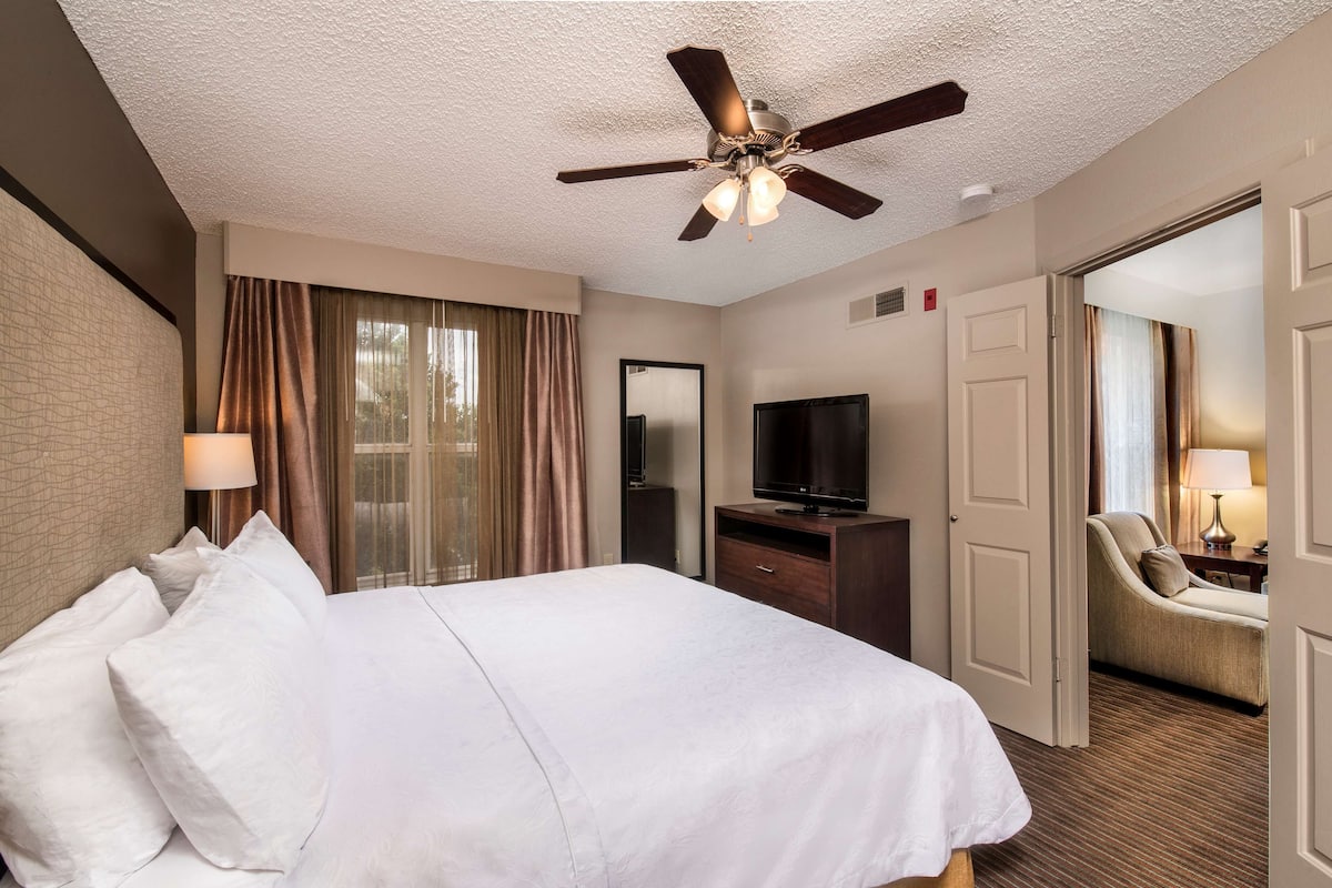 A hotel room with a large bed, a ceiling fan, a TV, a desk, and a chair.