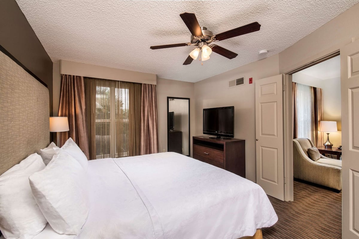 A hotel room with a bed, a television, a desk, a chair, and a ceiling fan.