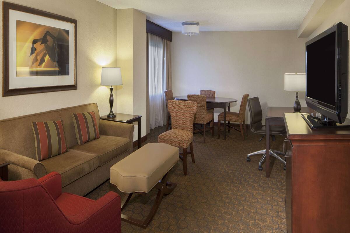 A hotel room with a sofa, a dining table with chairs, a TV, and a lamp.