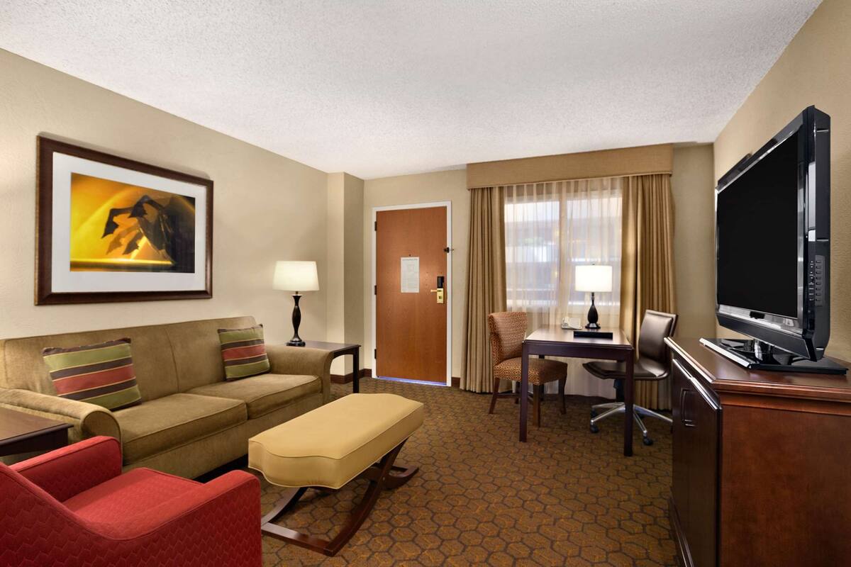 A hotel room with a sofa, armchair, ottoman, dining table, chairs, a television, and a painting on the wall.