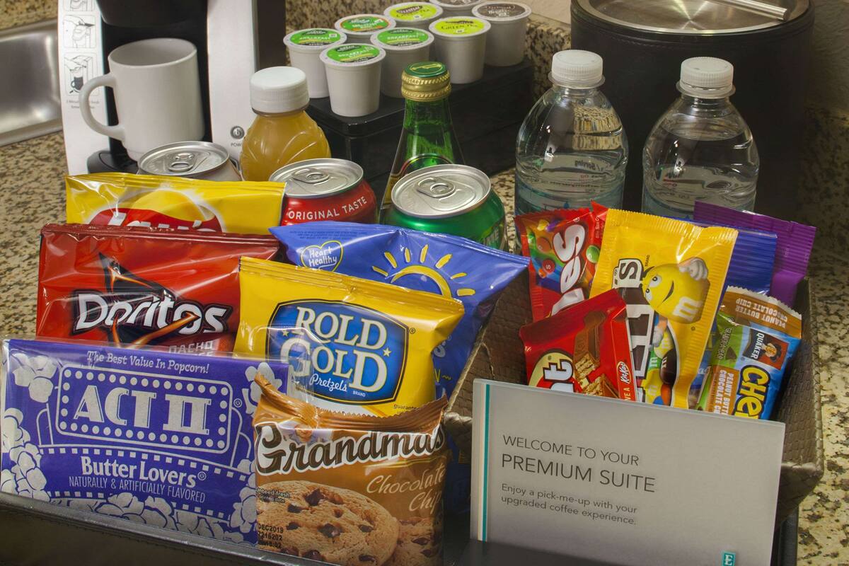 A breakfast buffet with a Keurig coffee maker, various beverages, snacks like Doritos, Rold Gold pretzels, and Butter Lovers cookies.