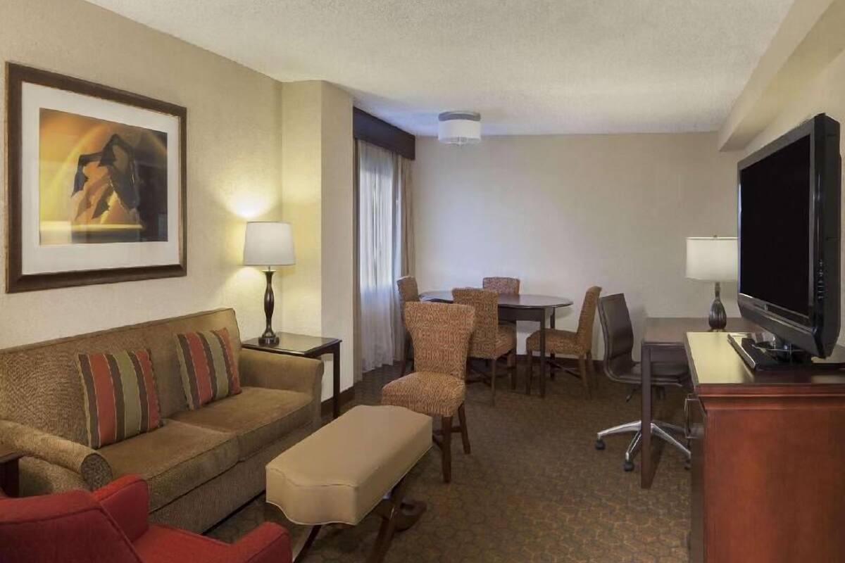 A hotel room with a sofa, armchair, dining table, chairs, a television, and a lamp.