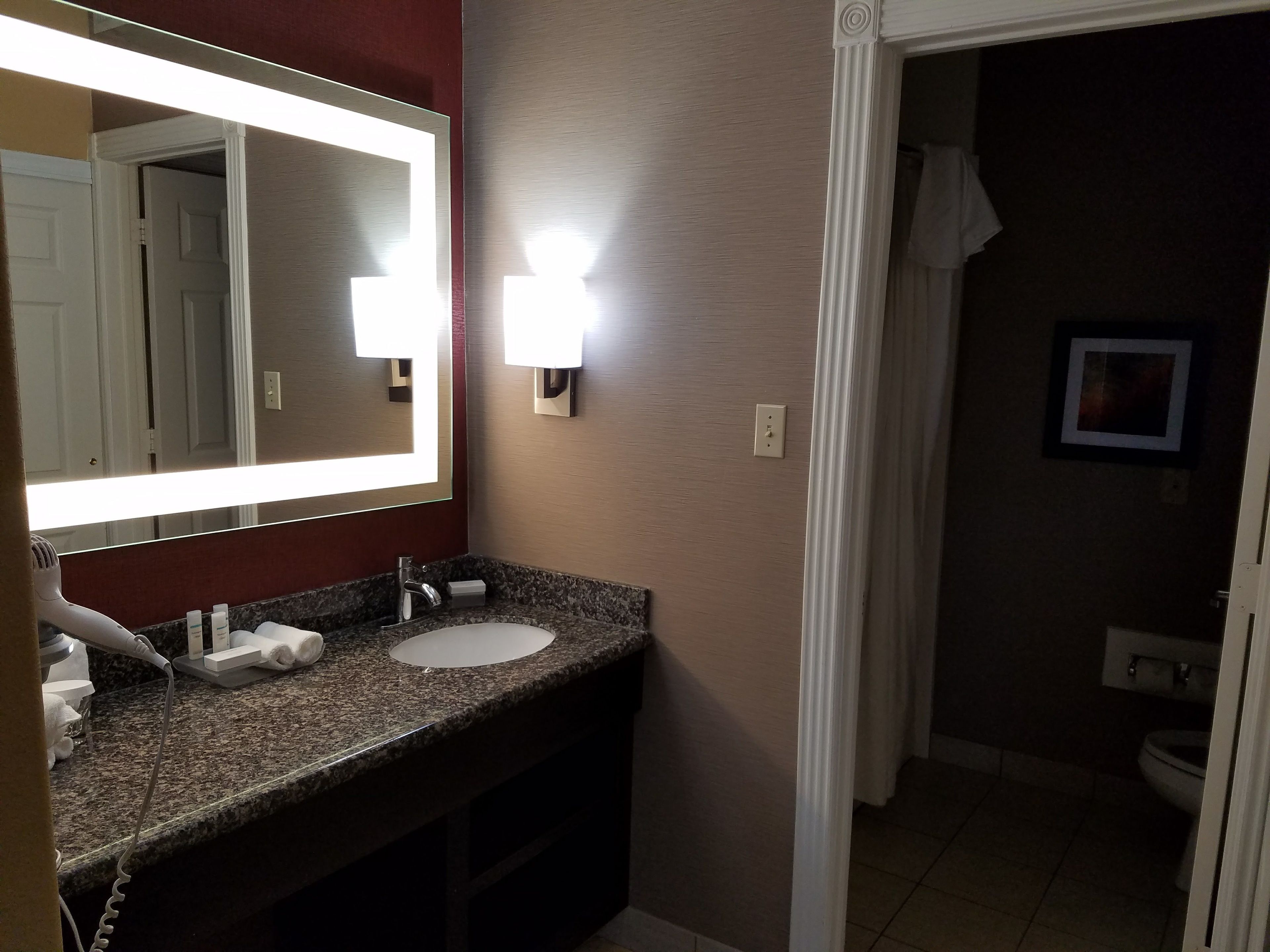 A bathroom with a granite countertop, a white sink, and a large mirror with lights. There is a towel rack and a framed picture on the wall.