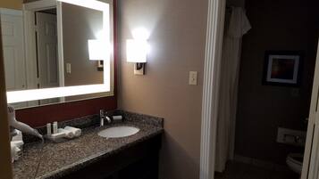 A bathroom with a granite countertop, a white sink, and a large mirror with lights. There is a towel rack and a framed picture on the wall.