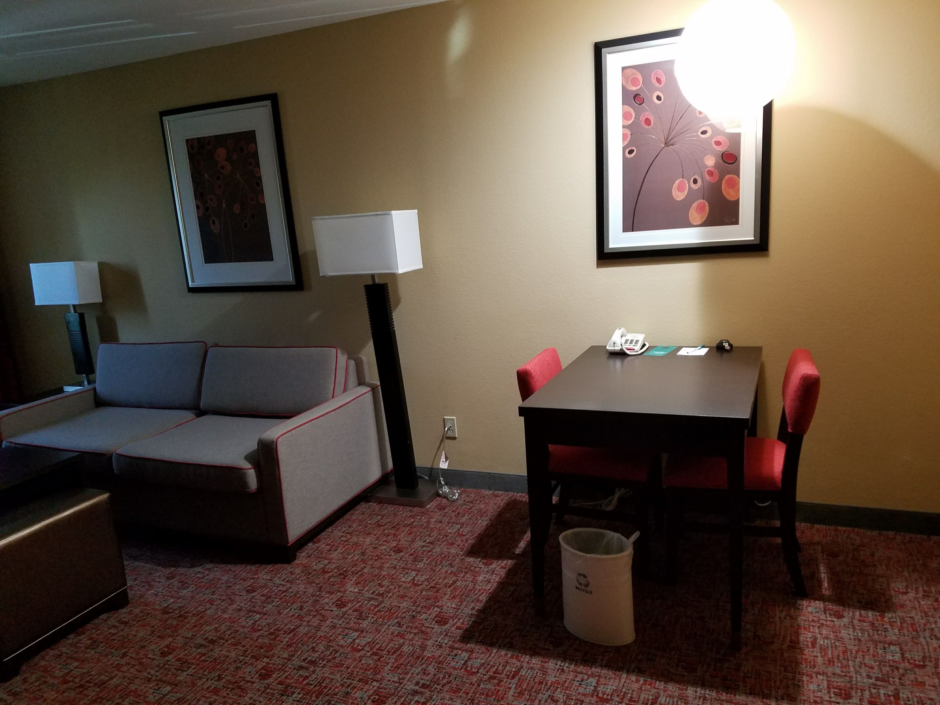 Habitación de hotel con sofá, una mesa de comedor con sillas, un cubo de basura y dos lámparas.
