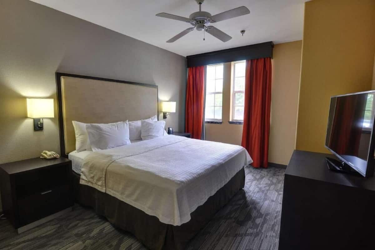 A hotel room with a bed, a nightstand with a phone, a TV, a ceiling fan, and a window with red curtains.
