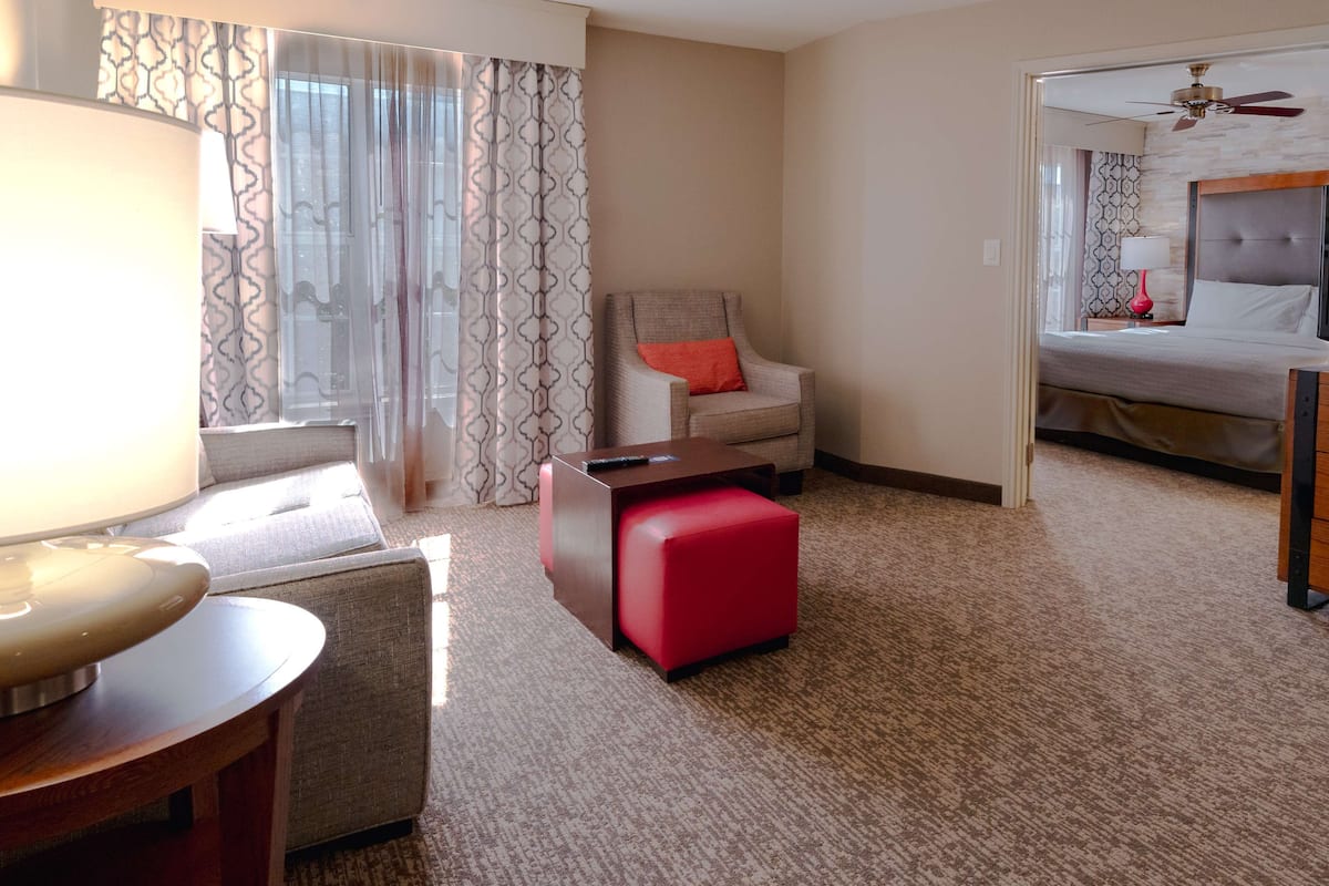Habitación de hotel con cama, un reposapiés rojo, una silla con cojín, un escritorio con lámpara y vistas al pasillo.