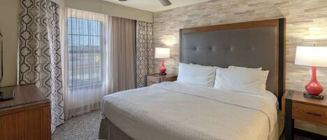 A hotel room with a large bed, a ceiling fan, a wooden nightstand, a red lamp, and a patterned curtain.