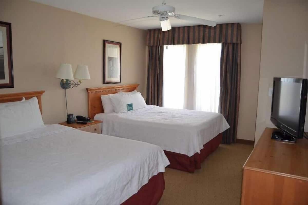 Habitación de hotel con dos camas, televisión, ventilador de techo y ventana con cortinas.