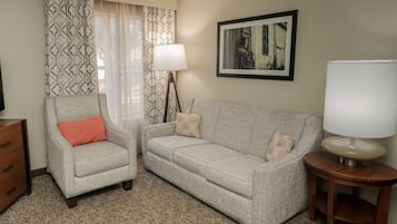 A living room with a grey sofa, a matching armchair, a wooden side table with a lamp, a floor lamp, and a framed picture on the wall.