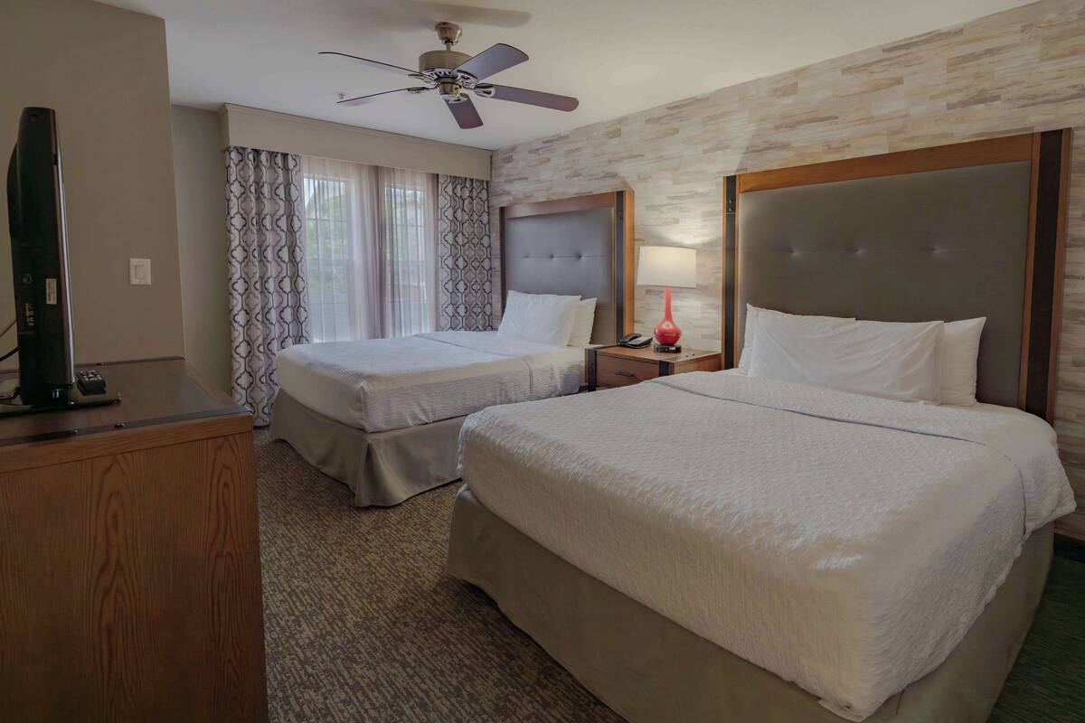 Una habitación de hotel con dos camas, un ventilador de techo, un televisor, una lámpara y un aparador de madera.
