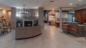 Un hall de hotel con un mostrador de recepción curvo, una chimenea, un candelabro y una zona de cocina al fondo.