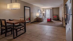 Una habitación de hotel con una mesa de comedor, sillas, un sofá, un reposapiés rojo, un refrigerador y un espejo.