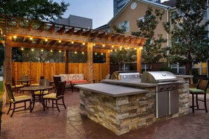 An outdoor patio with a stone barbecue station, wooden chairs, and a pergola with string lights. - Homewood Suites by Hilton Houston-Westchase (Houston)