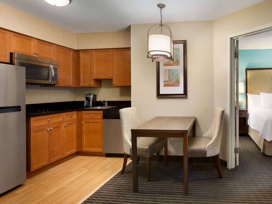 A compact hotel room with a kitchenette, a dining area with a table and chairs, a bed, and a framed artwork on the wall.