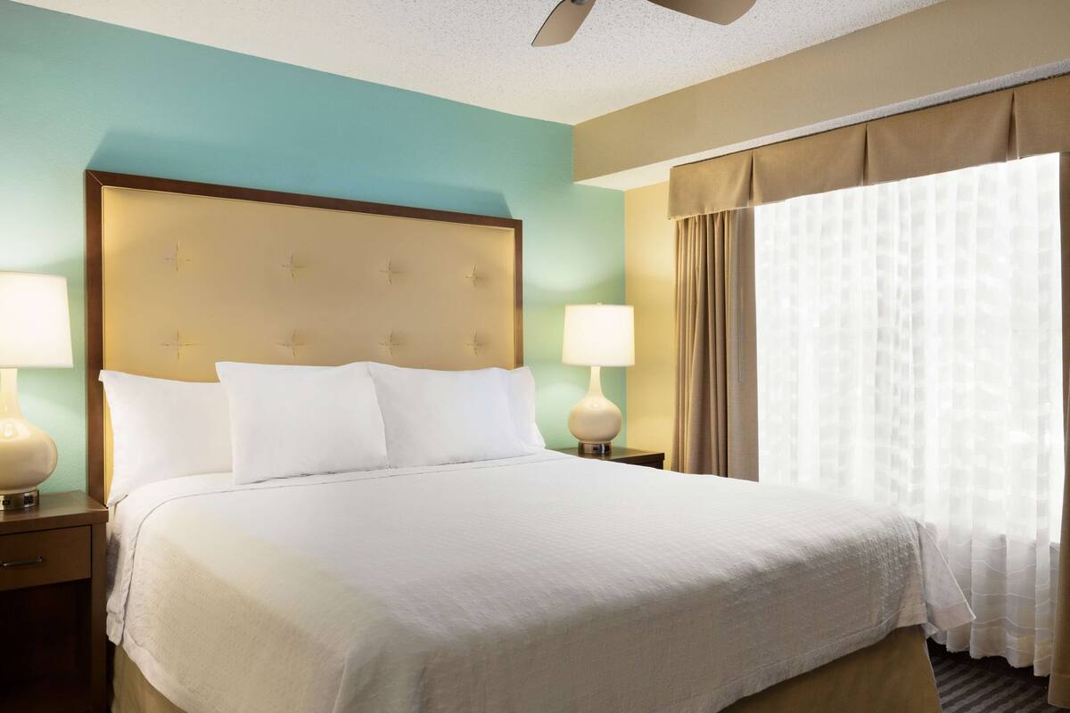 A hotel room with a large bed, two bedside tables with lamps, a ceiling fan, and a window with sheer curtains.