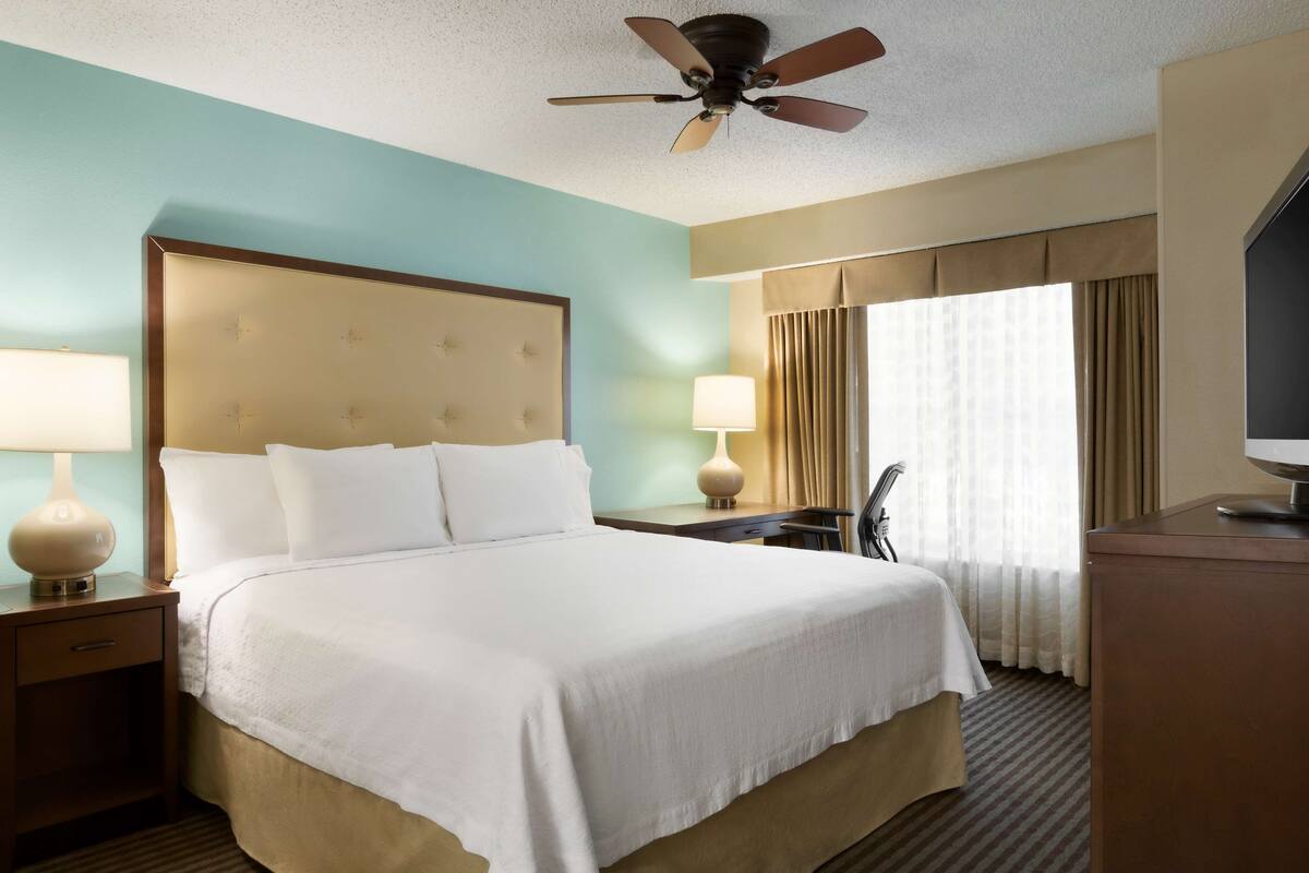 A hotel room with a large bed, a desk, a chair, a television, and a ceiling fan.
